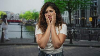 Fototapeta premium Hispanic woman with hands on cheeks, freckled face and white t shirt standing on street by canal in amsterdam; urban anxiety.