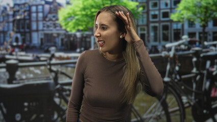 Fototapeta premium Woman young blonde with hand to hair beside bicycles along a canal on a street in amsterdam wearing brown top, gazing aside; quiet contemplation.