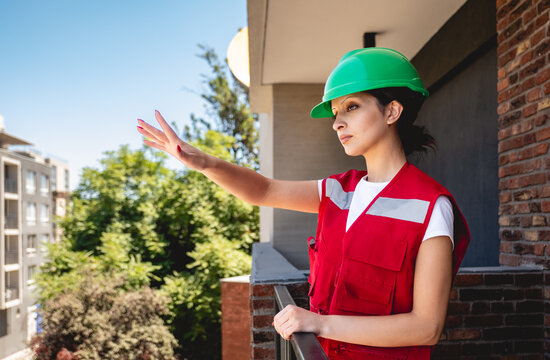 Young female safety inspector wearing green safety helmet and red safety vest, supervising construction site safety and giving instructions from balcony in sunny day