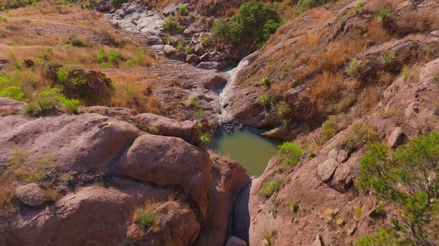 Aerial drone footage captures a rugged desert canyon with natural rock pools and turquoise water surrounded by dry golden grass, red boulders, and scattered green vegetation under warm sunlight.
