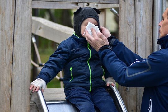 Parent wiping toddler&rsquo;s nose with tissue on playground slide, caring outdoor moment showing protection, hygiene and family support during cold weather in everyday life scene.