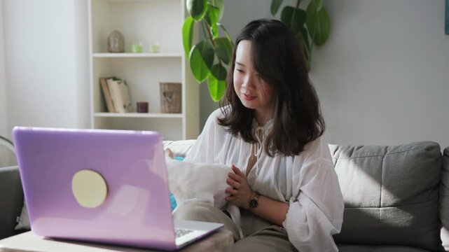 Pleasant-looking young Chinese woman holding small cute pet in hands. Fine girl using laptop, browsing. Modern apartment. City lifestyle. Inche. Weight. Indoors.