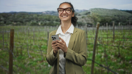 Naklejka premium Woman holding smartphone and smiling while typing with both hands in vineyard; contentment rural work.