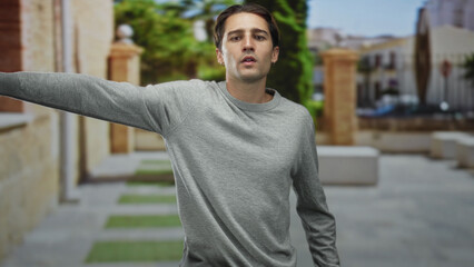 Fototapeta premium Man in gray sweater points finger and extends arm sideways on a sunlit stone street near benches and archway; curiosity decision.
