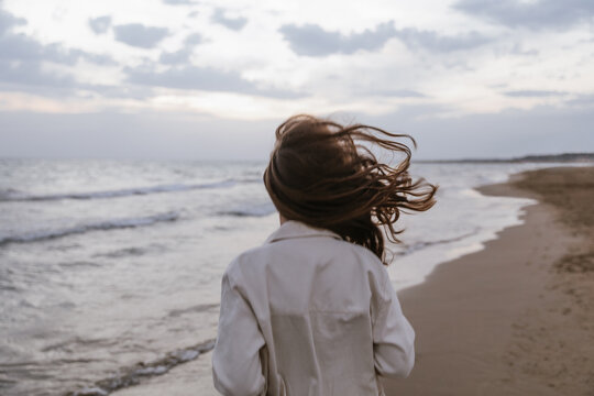 A cinematic rear view of a young woman standing on beach, her hair blowing wildly in the strong wind. The overcast sky and moody ocean waves create a feeling of peaceful solitude, raw nature, freedom