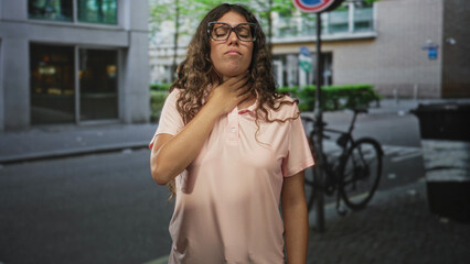 Obraz premium Hispanic teenage woman holding hand to neck and wearing glasses on a street near a parked bicycle and building entrance; fatigue discomfort.