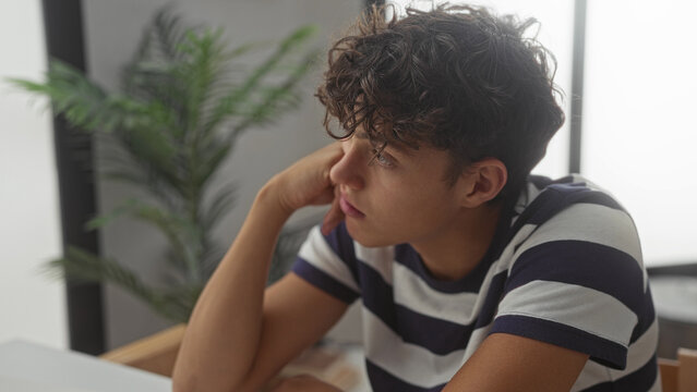 Teen boy with curly brown hair resting chin on hand, wearing striped shirt and leaning at table in studio; quiet pensive reflection.