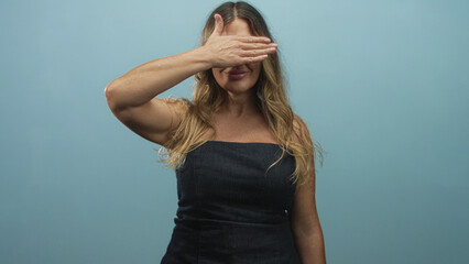 Fototapeta premium Woman wearing strapless denim dress smiling with hand covering eyes and exposed forearm in studio, eyeglasses visible in other frames; playful shyness.