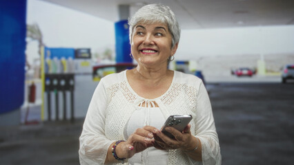 Woman tapping smartphone with hands at gas station forecourt, smiling while checking messages  happiness connection. © Krakenimages.com