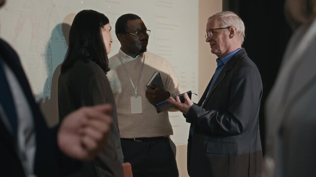 Rack focus shot of multiethnic conference participants with badges engaged in conversation in front of projection screen with unrecognizable business couple shaking hands in foreground