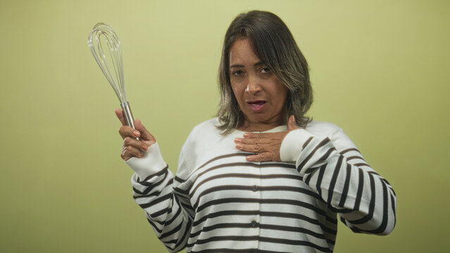 Woman holding whisk with hand on chest, striped sweater visible, expressive gasp gesture in studio; surprise.