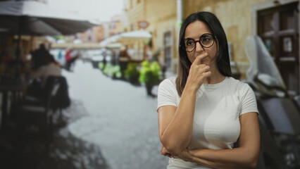 Fototapeta premium Woman in white tshirt with hand on chin and glasses, arms crossed, standing on a street beside cafe tables and scooter; thoughtful reflection.