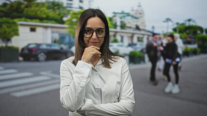 Fototapeta premium Woman in white shirt and glasses, palm up presenting with one hand then hand to chin pensive pose on an urban street crosswalk; thoughtful decision.