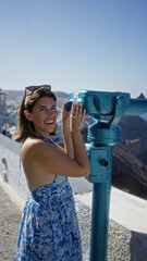 Obraz premium Woman looking through coin operated viewer on street in santorini town near caldera cliff and white buildings; curiosity travel.