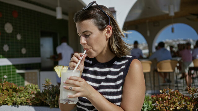 Woman sipping a frosty cocktail through a straw at a building bar counter with sunglasses on head and a pineapple garnish; relaxed vacation.