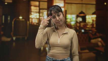 Fototapeta premium Young woman flicks fingers and rolls eyes, hand visible in a building lobby with luggage cart and pendant lights; subtle annoyance.