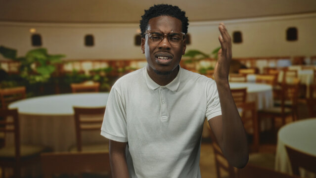 African american man wearing white polo shirt raising right hand gesture inside restaurant building; confusion.