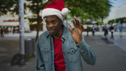 Obraz premium Man wearing santa hat and denim jacket waves hand with friendly smile on lively city street lined with trees; holiday joy.