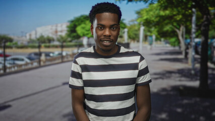 Young african american man with bare arms stands smiling in sunlit street beside parked cars and trees  confidence. © Krakenimages.com