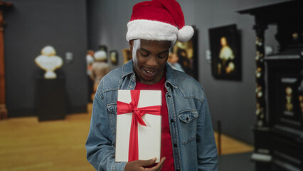 Obraz premium Man wearing santa hat and denim jacket holding white gift box wrapped in red ribbon and pointing finger in building; holiday joy.
