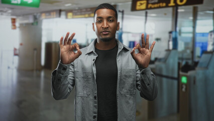 Fototapeta premium Man holding both hands in ok gesture at airport gate near boarding kiosks and gate 10 sign, wearing gray shirt and black tee; confidence.