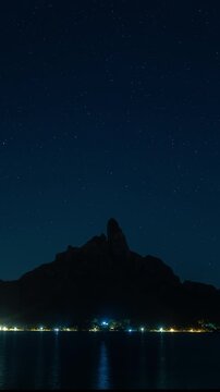 Vertical Screen: Mount Otemanu in Bora Bora rises dramatically against a starry night sky transitioning to dawn. Coastal lights reflect on calm lagoon waters as purple and blue hues paint the horizon.