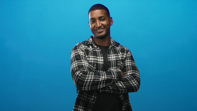 Young black man in black and white plaid flannel shirt with arms crossed, smiling in a studio blue backdrop; confidence.