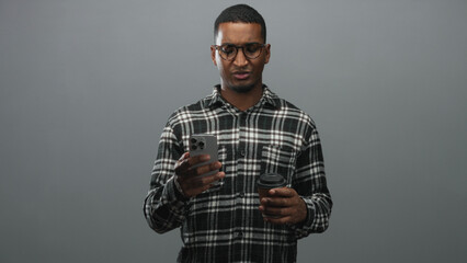 Fototapeta premium Black man wearing glasses and plaid shirt holding a smartphone in one hand and a takeaway coffee cup in the other, frowning while reading the screen in studio; confusion.