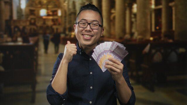 Young man holding indonesian rupiah banknotes and giving thumbs up in catholic church building; joy prosperity.
