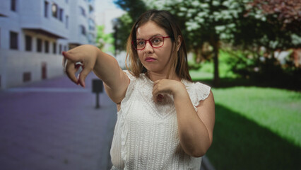 Woman extends arm and points finger on a street beside a building wearing red glasses and a white...