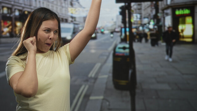 Woman on city street clasping hands and raising a fist with bare forearms visible, smiling while turning toward traffic and pedestrians; determination.