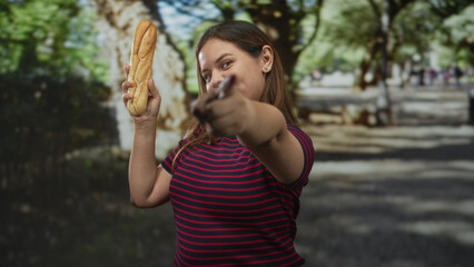 Obraz premium Woman holding baguette aloft with raised hand and smiling beside benches on a tree lined street under dappled sunlight; playful confidence.