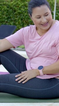 Vertical video: Smiling woman stretching on mat by pool responding to friend offscreen in pink top