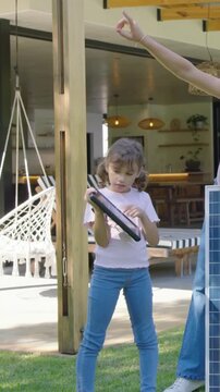 Vertical video: After mom pointing sun, kid in pink tee angling small solar panel by big panel yard
