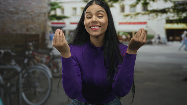 Woman in purple sweater pinches fingertips to lips in sunlit city street by parked bicycles; delight taste flavor satisfaction.