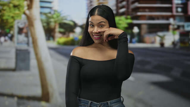 Young latin woman wearing offshoulder top and jeans pointing finger to nose on city street; curiosity.