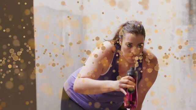 Female exerciser finishing set, sipping red bottle then bending and recovering under golden bokeh