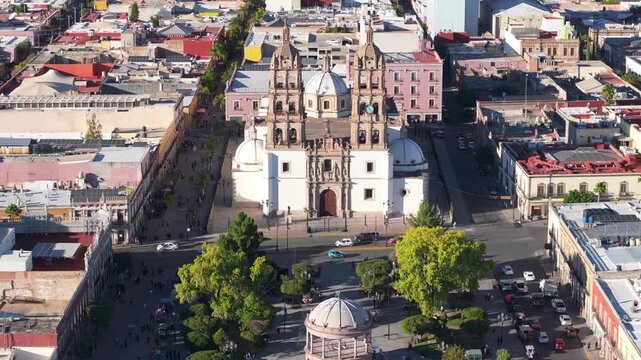 Drone footage ascending over ornate baroque cathedral in Durango Mexico reveals twin bell towers with clock and elaborate stone facade surrounded by historic downtown streets and colorful colonial.