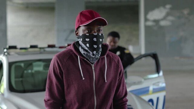 Portrait african american offender with hat in mask stand in patrol car in handcuffs burglar crime hold danger gangster gun robber handgun illegal law outlaw arrest bad guy close up slow motion