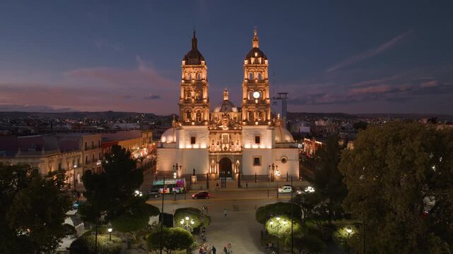 Aerial drone footage of the Cathedral Basilica of Immaculate Conception in Durango, Mexico at twilight, featuring illuminated baroque twin bell towers against a purple and orange sunset sky.