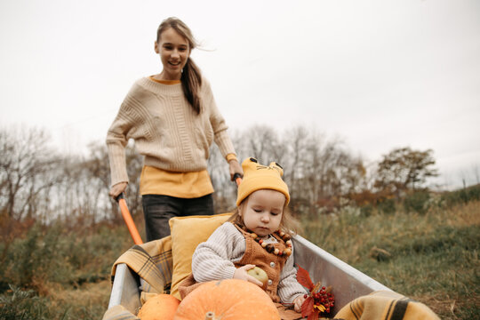 Sisters riding in wheelbarrow with pumpkins during autumn day