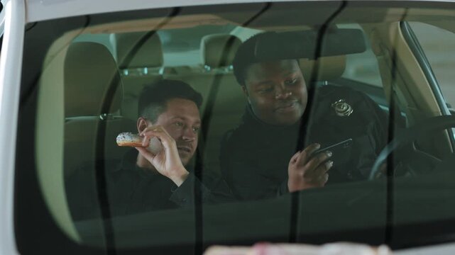 In patrol car two policemen eating donut use phone smile talk sitting help happy african american break rest gangster uniform funny teamwork portrait slow motion