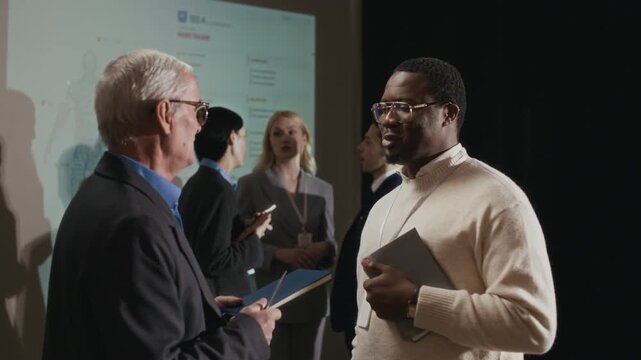 Zoom out rack focus shot of multiethnic businessmen discussing lecture in conference hall with three other participants in formalwear talking in front of projection screen in background
