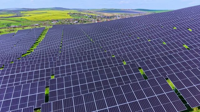 Rows of solar panels stretch across a green field in a rural area. Hills and small towns can be seen in the distance under a bright blue sky. Work on renewable energy is taking place.