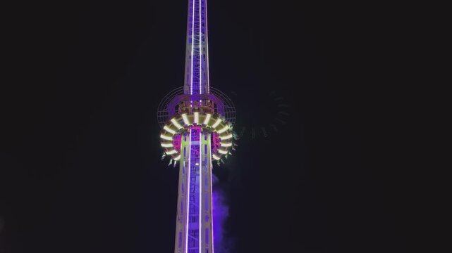 Thrilling amusement park drop tower ride illuminated with neon purple and white lights at night.