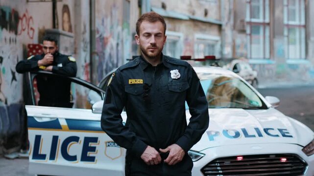 Attractive smiling young man cops stand near patrol car look at camera enforcement officer police uniform auto safety security communication control policeman close up slow motion