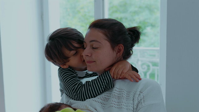 Young boy embracing his mother near window, wrapping arms around her neck with eyes closed, tender expression of love, safety, and emotional closeness at home