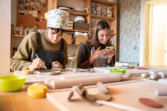 Women painting clay piece and using phone in pottery workshop studio