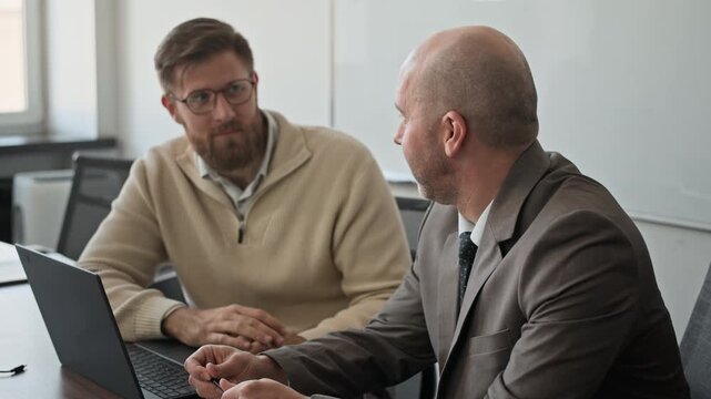 Tilt shot of two Caucasian colleagues sitting at wooden desk and chatting. They turning heads to camera and smiling lightly