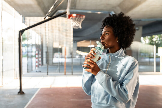 Young woman athlete drinking water at an outdoor basketball court